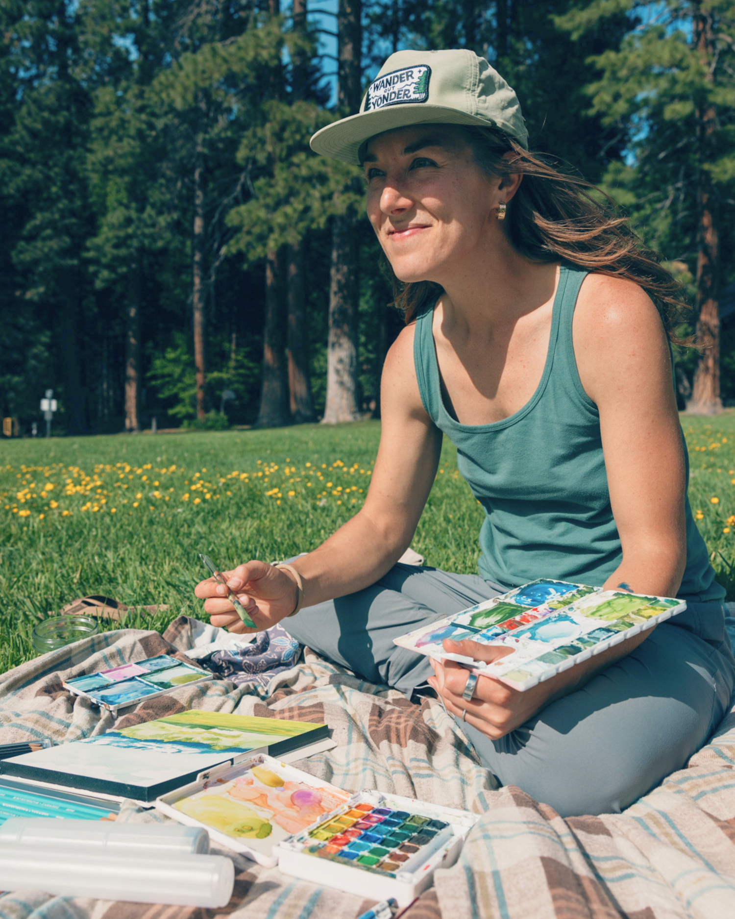 The landscape painter, Maria Miller is sitting on a blanket outdoors painting with art supplies, surrounded by nature.