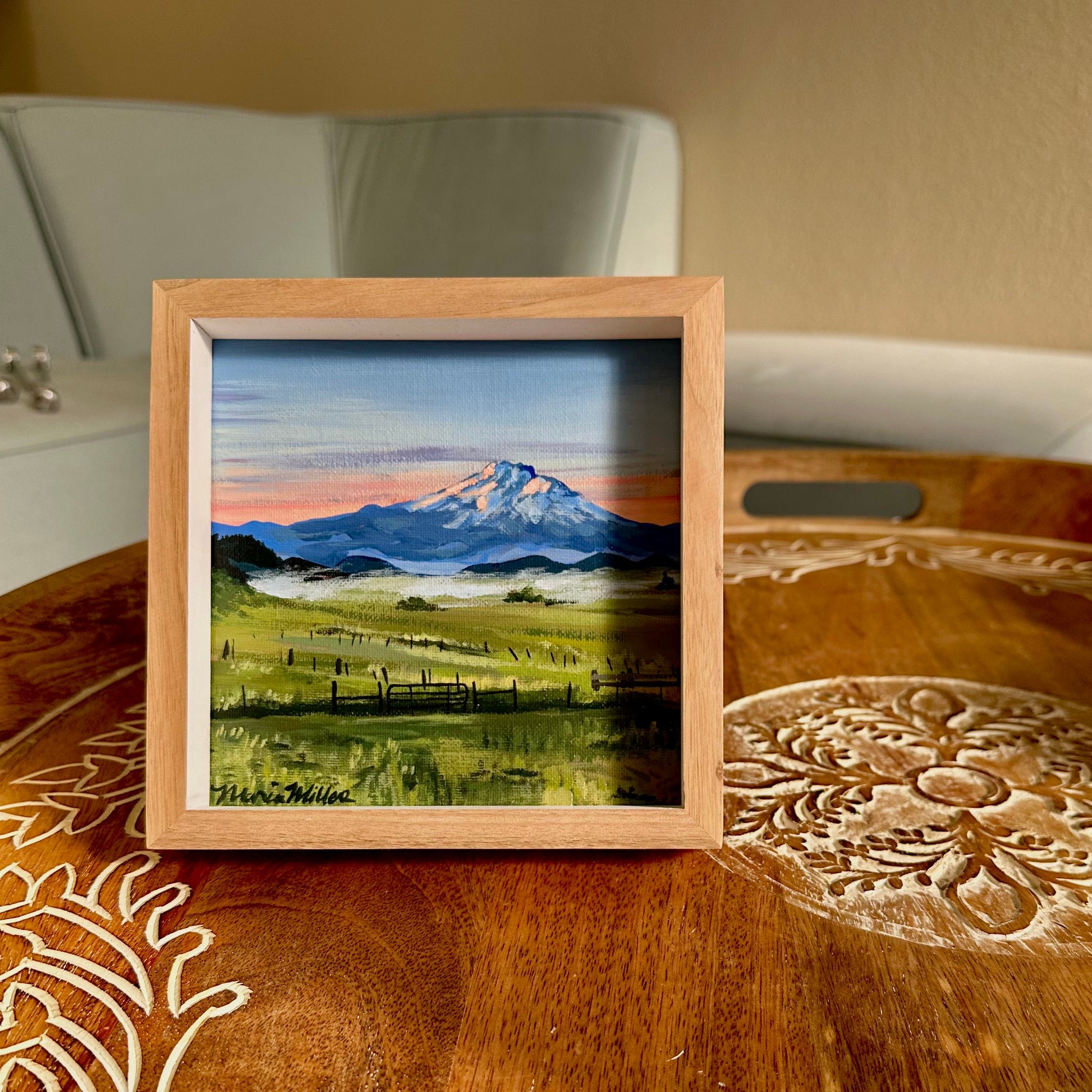 A small square framed landscape painting of a snow-capped mountain at sunrise, displayed on a carved wooden table.